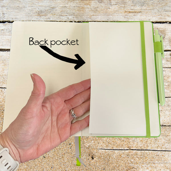Hand holding a notebook with a back pocket labeled and an arrow pointing to it on a wooden surface.